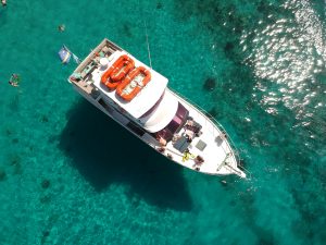 Aerial view of a white catamaran anchored in shallow, crystal-clear turquoise water with people snorkeling around it on a sunny day.