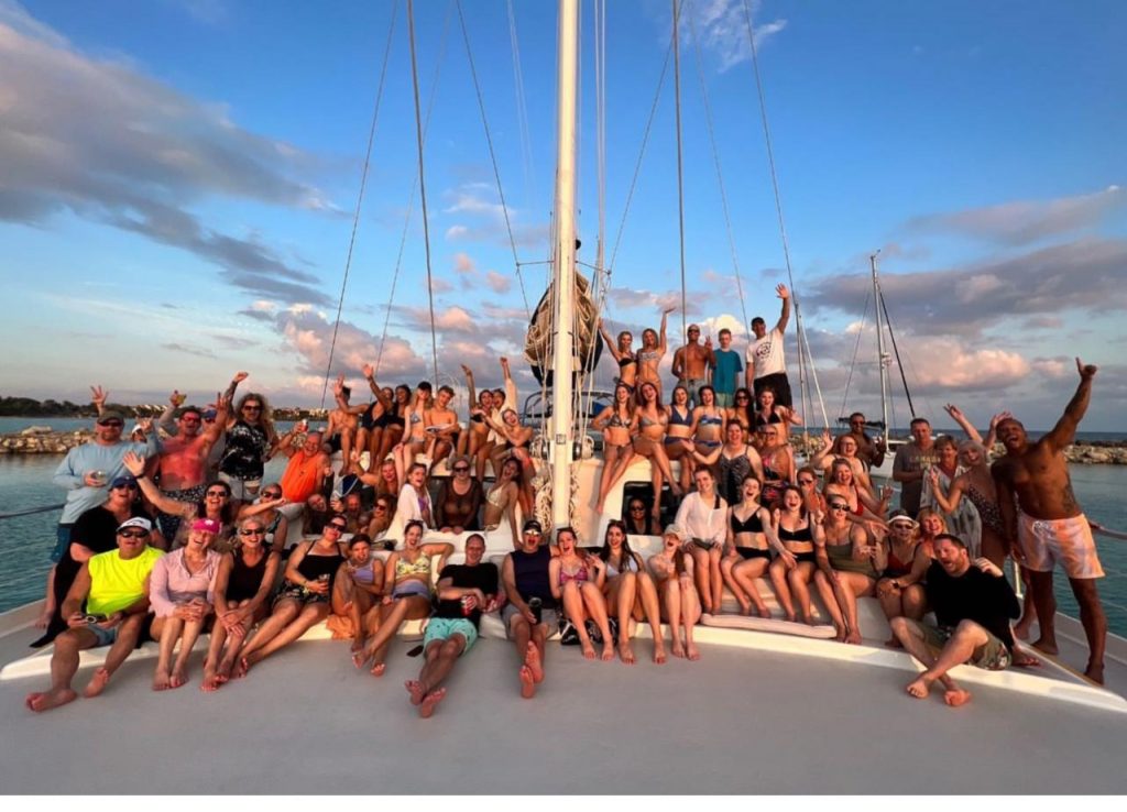 Large group of adults posing together on the deck of a sailboat at sunset, smiling, raising their arms, and celebrating while anchored near a rocky shoreline under a colorful sky.