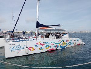White catamaran named “La Trinidad” with colorful fish illustrations along the hull, carrying a group of passengers seated under a shaded canopy while cruising near a coastal city skyline.